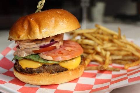 Gourmet burger with multiple meat patties, cheese, tomato, and onion on toasted bun served with french fries on checkered paper