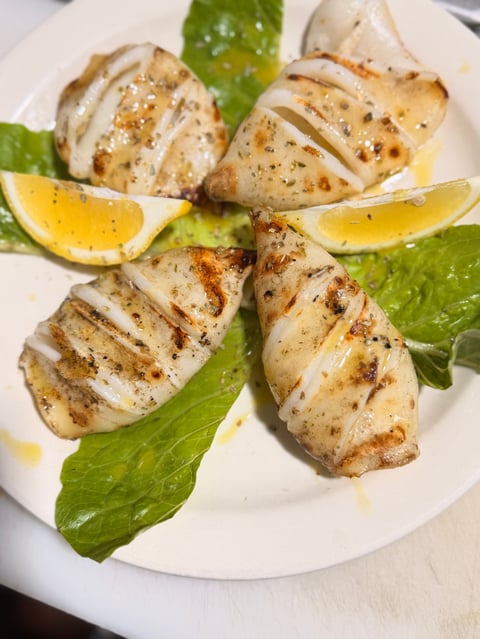 Four grilled squid tubes with char marks, arranged on a white plate with fresh basil leaves and lemon wedges